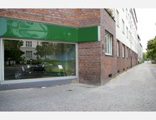 Backsteingebäude mit großer Schaufensterfläche, hinter der ein schwarzes Auto und grüne Pflanzen erkennbar sind. Im Vordergrund verläuft ein gepflasterter Gehweg entlang der Gebäudefassade.