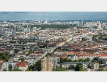 Luftaufnahme einer städtischen Landschaft in Berlin mit einer Mischung aus modernen und historischen Gebäuden. Im Hintergrund ragt der Berliner Fernsehturm über die Stadt hinaus, umgeben von einer Vielzahl dicht stehender Häuser und grünen Parkflächen.