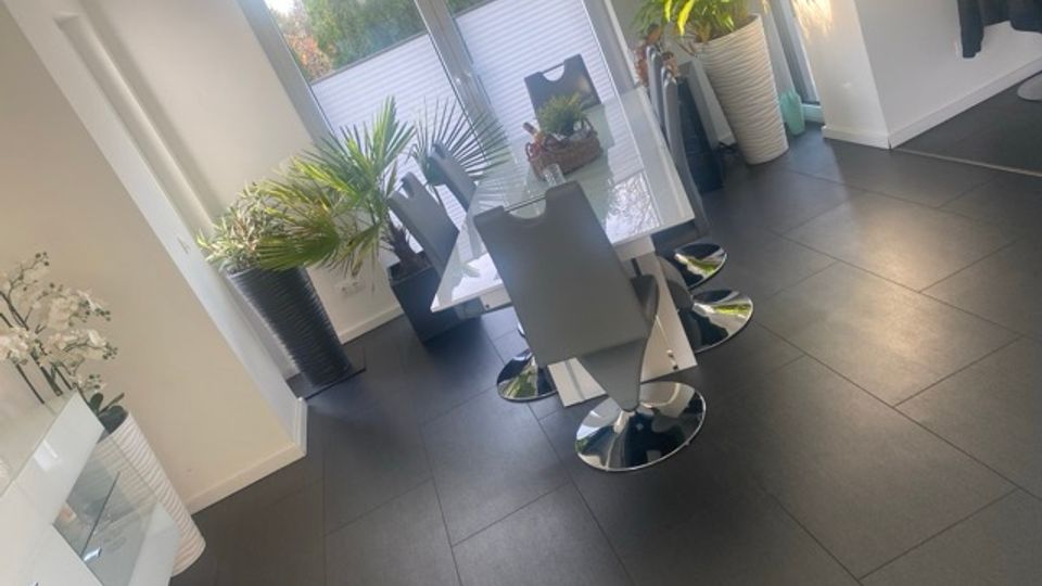 Heller moderner Essbereich mit dunkelgrauen Fliesen, einem weißen Esstisch und mehreren grauen Stühlen mit Chromfuß. Große Glastüren führen auf eine Terrasse; mehrere große Zimmerpflanzen in hohen Töpfen stehen entlang der Fensterfront.