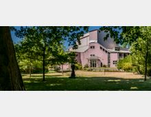 Ein modernes Gebäude mit lila Fassade und schrägen Dachflächen, umgeben von einem parkähnlichen Garten mit Bäumen, Rasen und einer Skulptur im Vordergrund. Ein gläserner Eingangsbereich ist zentral sichtbar, eingerahmt von Blumenbeeten und dichter Vegetation.