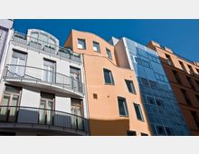 Mehrstöckige Wohngebäude mit modernen Fassaden, darunter weiße und orangefarbene Putzfassaden mit Balkonen sowie eine gläserne Fensterfront; klares Wetter mit blauem Himmel.