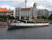 Ein Ausflugsschiff mit der Aufschrift "Stralau" fährt auf einem Fluss, im Hintergrund moderne Gebäude mit roten Dächern und historischen Elementen sowie der Berliner Fernsehturm. Der Himmel ist teils bewölkt, und am Ufer stehen Bäume.
