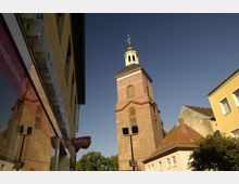 Straßenszene mit einem historischen Kirchturm aus Backstein und hellem Putzelementen, umgeben von Wohn- und Geschäftshäusern. Im Vordergrund spiegelt sich der Turm in der Glasfassade eines Gebäudes.