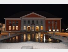 Ein Kulturzentrum in einem symmetrischen Gebäude mit orangener Fassade und weißem Säuleneingang, beleuchtet bei Nacht. Davor befinden sich ein Wasserbecken mit kleinen fontänenartigen Wasserspielen sowie Sitzbänke auf einem gepflasterten Platz.