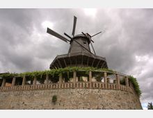 Historische Windmühle aus Holz auf einem erhöhten gemauerten Sockel mit umlaufender Pergola, die von Kletterpflanzen bewachsen ist, unter einem bewölkten Himmel.