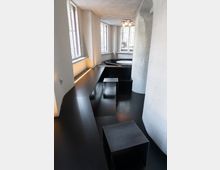 Moderner Innenraum mit glatten, schwarzen Sitzflächen und Tischen, großen Fenstern mit Gitterstäben und grob verputzten, weißen Wänden. Ein runder Deckenleuchter sorgt für Beleuchtung in der minimalistischen Gestaltung.