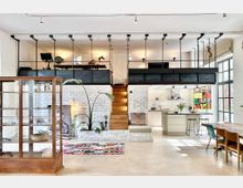 Großzügiger, offener Wohnraum in einem industriellen Stil mit weiß gestrichenen Ziegelwänden und einer hölzernen Treppe, die zu einer Galerie-Ebene führt. Im Vordergrund steht eine Glasvitrine, daneben befinden sich ein bunter Teppich, moderne Möbel und eine helle Küche mit einer zentralen Kochinsel im Hintergrund.