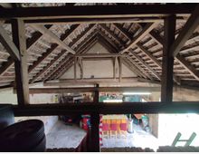 Dachboden eines Gebäudes mit sichtbaren Holzbalken und Ziegeldachkonstruktion, darunter ein offener Raum mit verschiedenen Möbeln, Lagergegenständen und Dekorationen. Rechts fällt Tageslicht durch eine große Öffnung ein.