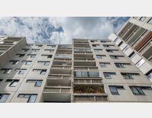 Mehrstöckiges Wohngebäude in Plattenbauweise mit Balkonen und rechteckigen Fenstern, aufgenommen aus einer Perspektive von unten gegen den Himmel. Im Vordergrund sind teils bepflanzte Balkone und kleine architektonische Details wie Beton-Geländer zu sehen.