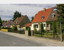 Reihe von Einfamilienhäusern mit Satteldächern und Vorgärten entlang einer gepflasterten Straße, umgeben von Bäumen und Zäunen aus Metall und Holz. Die Häuser haben verschiedene Dachfarben und teils verputzte, teils bewachsene Fassaden.