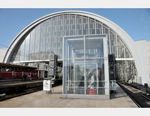 Bahnhof mit moderner Glas- und Stahlarchitektur, gekennzeichnet durch eine große, halbrunde Glasfassade im Hintergrund. Im Vordergrund befindet sich ein gläserner Aufzug, daneben Bahnsteige und ein roter Regionalzug.