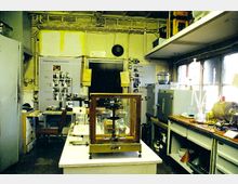 Ein kleines, älteres Labor mit verschiedenen wissenschaftlichen Geräten, darunter ein Holzrahmen mit einer Glasvitrine in der Mitte. An den Wänden hängen Poster und wissenschaftliche Diagramme, während Regale und Tische mit Equipment und Messinstrumenten gefüllt sind.