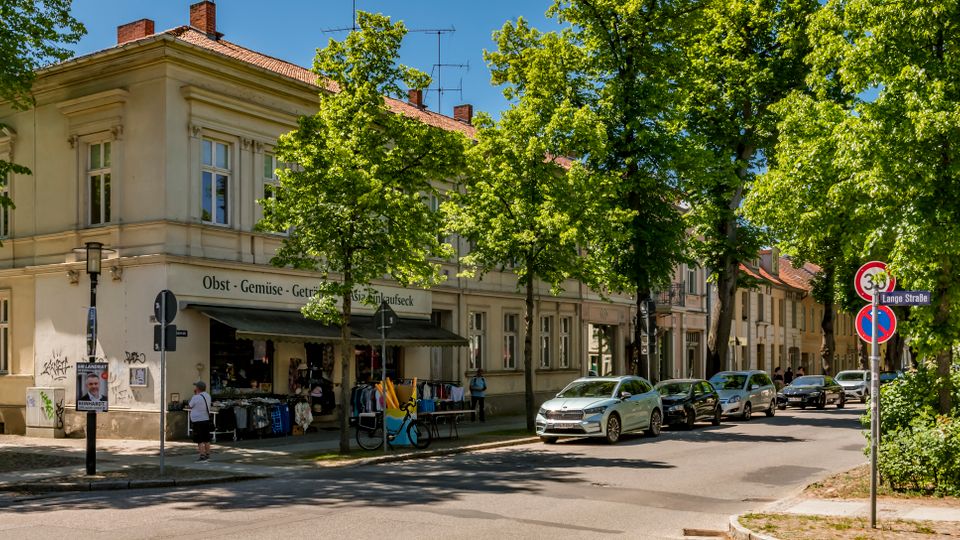 Straßenecke mit zweigeschossigem Altbau in heller Fassade und rotem Ziegeldach, im Erdgeschoss ein kleiner Laden mit Vordach. Entlang der von großen, belaubten Bäumen gesäumten Straße stehen parkende Autos und Verkehrsschilder am Gehweg.