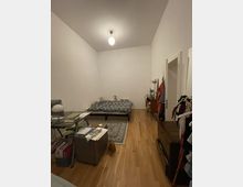 Ein einfach eingerichtetes Schlafzimmer mit einem Holzboden, einem Bett mit gemustertem Bettbezug, einem kleinen Teppich davor und einer Deckenlampe. Rechts befinden sich eine Kommode, einige aufgehängte Kleidungsstücke, und links ein Schreibtisch sowie Regale mit Büchern und Unterlagen.