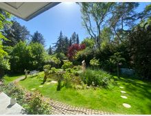 Gepflegter Garten mit kleinem Teich, Schilf und Trittsteinen im Rasen, umgeben von dichten Sträuchern und hohen Bäumen. Links führen ein gepflasterter Weg und eine Sitzbank durch die Grünfläche.