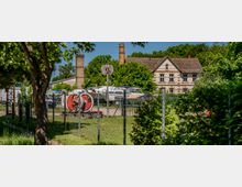 Eingezäuntes Industrie- und Lagergelände mit Booten und Geräten im Freien, im Hintergrund zwei hohe Backsteinschornsteine. Rechts steht ein zweigeschossiges, historisch wirkendes Gebäude mit hellem Putz, Ziermauerwerk und Satteldach, umgeben von dichtem Grün.
