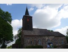 Historische Dorfkirche aus Feldstein mit einem Holzturm und spitzem Dach, umgeben von Bäumen und blauem Himmel mit weißen Wolken; vor der Kirche steht ein Schaukasten.