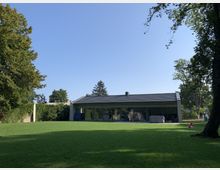 Modernes einstöckiges Gebäude mit Flachdach, großen Glasfronten und angrenzender Terrasse, umgeben von einem weitläufigen Garten mit gepflegtem Rasen und hohen Bäumen. Der Himmel ist klar und blau.
