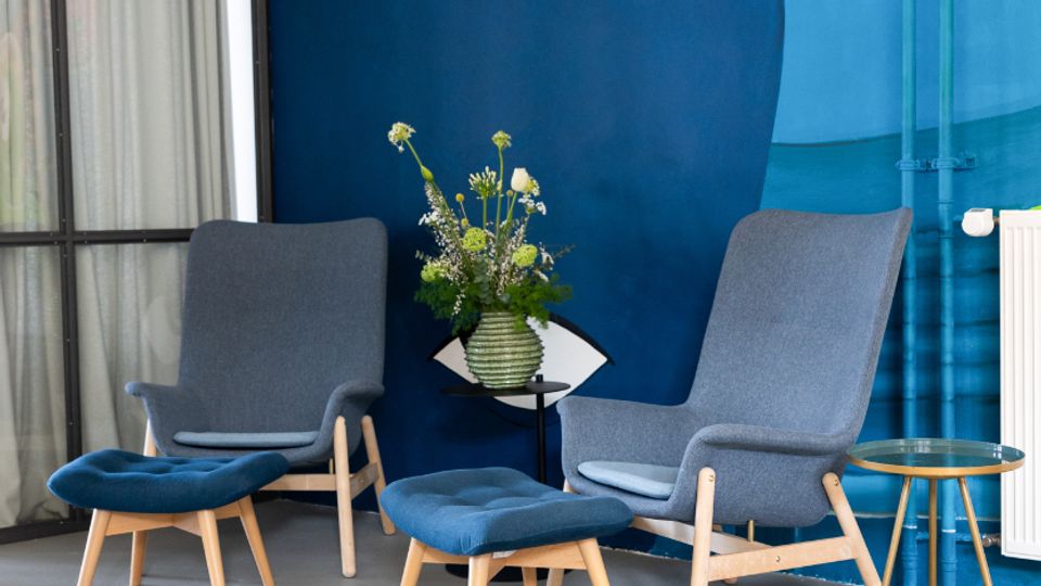 Sitzecke in einem modernen Innenraum mit zwei grauen Lounge-Sesseln, passenden Fußhockern und kleinen Beistelltischen auf glattem Boden. Im Hintergrund dominiert eine großflächige, farbenfrohe Wandmalerei mit abstraktem Gesichtsmotiv sowie ein Fenster mit Vorhang und ein Heizkörper.