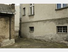 Ein schmaler Hinterhof mit zwei Gebäudewänden aus verputztem Mauerwerk, teils mit sichtbaren Schäden und abblätterndem Putz. Links ist ein kleines Gebäude mit Dachziegeln, rechts eine größere Wand mit vergitterten Fenstern auf verschiedenen Ebenen.