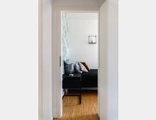 Blick durch eine geöffnete Tür in ein Schlafzimmer mit Holzfußboden, einem Bett mit dunkler Bettwäsche und Kissen sowie einer Nachttischlampe auf einem schwarzen Nachttisch. An der Wand hinter dem Bett befindet sich grüne Tapete mit Muster und ein kleiner Wandrahmen mit Vase.