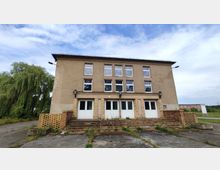 Ein dreistöckiges Gebäude mit einfacher Putzfassade und großen rechteckigen Fenstern, flankiert von zwei Laternen an der Front. Der Eingangsbereich besteht aus mehreren weißen Türen, erreichbar über eine kurze Treppe, umgeben von niedrigen Ziegelmauern; im Hintergrund sind Grünelemente und ein bedeckter Himmel sichtbar.