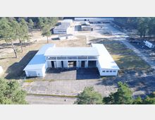 Luftaufnahme eines industriellen Gebäudekomplexes mit einem zentralen, großen weißen Gebäude mit Flachdach und mehreren Garagentoren im Vordergrund, umgeben von weiteren Werkshallen und angrenzendem Gelände. Um die Anlage befinden sich lichte Waldflächen mit Kiefern und vereinzelten Zufahrtswegen.