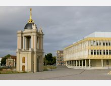 Ein Platz mit einem barocken Glockenturm links, verziert mit Statuen und goldener Spitze, und einem modernen gelben Gebäude rechts mit rechteckiger Fassade und Säulen. Im Hintergrund sind Bäume und weitere Gebäude zu sehen.