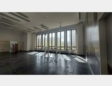 Großer, heller Innenraum mit hohen Fenstern, die den Blick auf eine städtische Umgebung bieten. Der Raum hat einen schwarzen Steinboden, weiße Wände und Decken mit Leuchtstofflampen, sowie einige Stehtische vor den Fenstern.