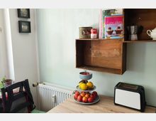 Eine helle Küchenecke mit einem Holzregal an der Wand, das mit Büchern, einer Kaffeemaschine und Dosen dekoriert ist. Auf dem Holztisch steht ein dreistufiger Obstständer mit Äpfeln, Zitronen und Tomaten sowie ein weiß-schwarzer Toaster.