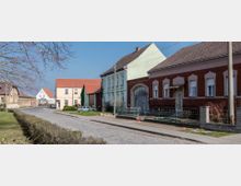 Straßenszene in einem Dorf mit Kopfsteinpflaster, flankiert von Häusern mit bunten Fassaden, darunter ein rotes Ziegelhaus und ein pastellgrünes Gebäude. Im Hintergrund sind weitere niedrige Gebäude und ein blauer Himmel sichtbar.