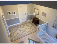 Treppenhaus mit hellem Holzfußboden und geometrischem Teppich. Wände mit weißer Zierleiste und grauer Tapete, ergänzt durch eine weiße Tür, gerahmte Bilder, eine kleine Kommode und ein Geländer aus weißen Streben.