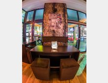 Innenraum eines modern gestalteten Cafés oder Restaurants mit einem dunklen Holztisch, zwei gepolsterten Hockern und einer dekorativen Tischleuchte. Im Hintergrund ist eine Wand mit abstrakter Musterung und einem Schwarz-Weiß-Porträt, daneben große Fenster mit Blick auf eine belebte Straße.