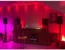 Ein Raum mit einer Musikanlage, bestehend aus einem hölzernen Sideboard mit Schallplattenspielern, Lautsprechern auf Ständern und indirekter Beleuchtung in Rot und Magenta. An der Wand ist ein Kunstbild angebracht, und der Boden weist eine moderne Möblierung mit einem Metallstuhl auf.