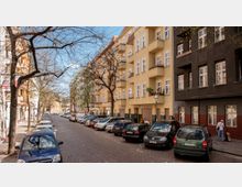 Straßenansicht mit Mehrfamilienhäusern im Altbaustil; links ein beige-gelbes Gebäude mit Balkonen, rechts ein dunkelbraunes Haus. Autos parken entlang der gepflasterten Straße, die von kahlen Bäumen gesäumt ist. Im Hintergrund ist ein sandfarbenes Gebäude zu erkennen.