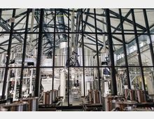 Innenansicht einer modernen Brauerei mit großen, silbernen Edelstahlbehältern und komplexen Rohrleitungen. Die Struktur des Gebäudes zeigt ein Glasdach mit schwarzen Stahlträgern, und die Ausstattung im Vordergrund besteht aus mehreren kleineren Behältern.