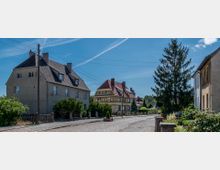 Straßenszene in einem Dorf mit gepflastertem Weg, umgeben von mehreren zweigeschossigen Wohnhäusern, darunter ein Fachwerkhaus mit rotem Dach. Im Vordergrund befinden sich Bäume, Sträucher und ein großer Nadelbaum, während der Himmel klar und blau ist.