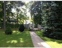 Gartenanlage mit gepflastertem Weg, umgeben von Bäumen und Sträuchern, die zu einem Eingang mit Steintreppe und schmiedeeisernem Gittertor führen; im Hintergrund stehen moderne Gebäude.