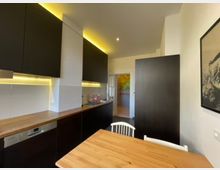 Moderne Küche mit dunklen Schränken, Holzarbeitsplatten und warmweißer Unterbeleuchtung. Im Vordergrund ein Holztisch mit weißen Stühlen, im Hintergrund ein Durchgang zu einem weiteren Raum mit einem bunten Wandbild.