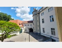 Innenhof eines historischen Gebäudekomplexes mit weiß verputzten Fassaden, einem runden Turm mit Fenstern und einem Torbogen. Im Vordergrund ein gepflasterter Platz, ein Baum und eine Laterne; im Hintergrund ein Gebäude mit roten Ziegeldach.