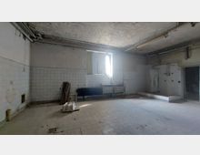 Ein leerer, heruntergekommener Raum mit weißen Fliesenwänden und einer gefliesten Bodenfläche. An der Wand befinden sich ein Fenster mit geöffnetem Holzrahmen, ein Heizkörper und eine Metallspüle; Rohrleitungen verlaufen entlang der Decke, während diverse Gegenstände auf dem Boden verteilt liegen.