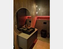 Modernes Badezimmer mit dunkler Wandverkleidung aus Holzlamellen und Akzenten in Betonoptik. Ein runder Wandspiegel, ein Waschtisch mit schwarzen Aufsatzwaschbecken und kupferfarbenen Armaturen, eine schwarze Toilette sowie rote indirekte Beleuchtung und drei Hängeleuchten bilden die Hauptmerkmale des Raums.