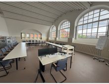 Heller Seminarraum mit U-förmig angeordneten Tischen und gepolsterten Stühlen, großen Rundbogenfenstern mit Blick nach draußen, weißer Decke mit Querbalken und einem Flipchart auf der rechten Seite. Im Vordergrund stehen ein Schreibtisch mit Monitoren und eine Tastatur.