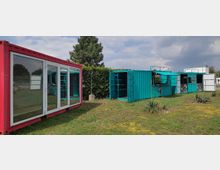 Zwei umgebaute Schiffscontainer auf einer Rasenfläche: Ein roter Container mit großen Glasfronten links und ein blauer Container mit geöffneten Türen und Klappen rechts. Im Hintergrund sind eine Hecke, ein Baum und ein bewölkter Himmel zu sehen.