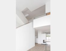 Heller, moderner Innenraum mit hohen Decken, weißen Wänden und Holzboden. Ein offener Kamin und bodentiefe Fenster sind sichtbar, mit Blick auf den Außenbereich.