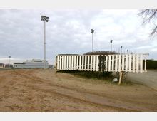 Offene Fläche mit sandigem Boden, einem weißen Holzzaun und einer Hecke im Vordergrund. Im Hintergrund sind Flutlichtmasten und ein modernes, mehrstöckiges Gebäude erkennbar, wahrscheinlich eine Sport- oder Veranstaltungsstätte.