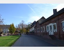 Straße mit Blick auf ein Ensemble aus historischen Backsteingebäuden und modernen Wohnhäusern, flankiert von Bäumen und einer Grünfläche mit Sitzbereichen und einem großen Tongefäß als Dekoration. Der Himmel ist klar und blau.