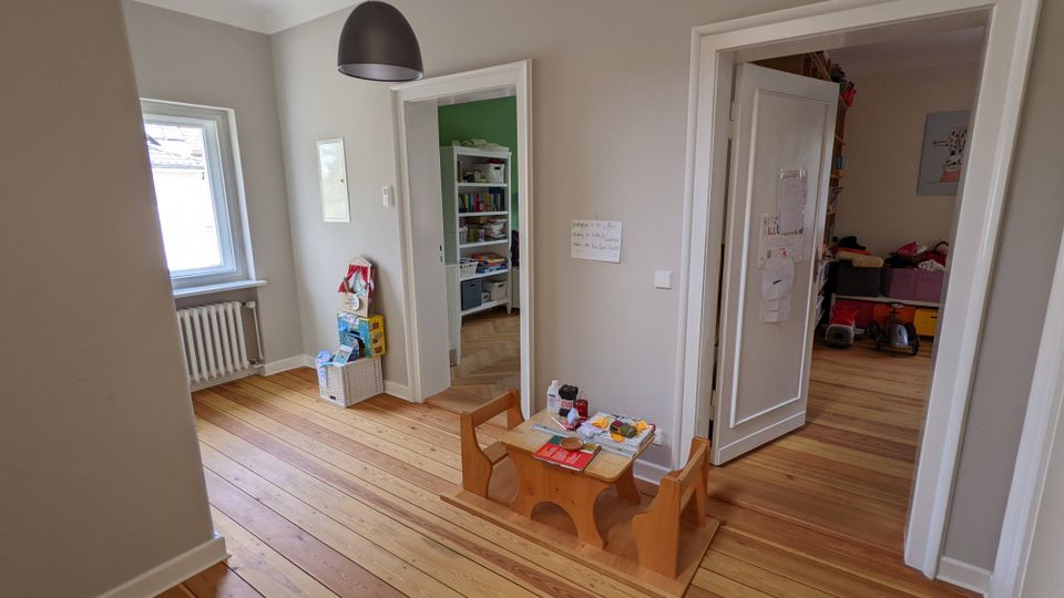 Heller Wohnungsflur mit breiten Holz-Dielen, grauen Wänden und schwarzer Pendelleuchte; links ein Fenster über einem Heizkörper. Zwei weiße Türrahmen führen in angrenzende Räume, im Vordergrund steht ein niedriger Kindertisch mit Bänken.