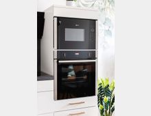 Weißer Einbauschrank in einer modernen Küche mit einem schwarzen Mikrowellen- und Backofenset aus Edelstahl, flankiert von grüner Dekoration im Vordergrund und einer mit Pflanzenmotiv gestalteten Wand im Hintergrund.