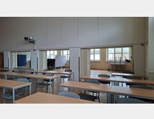 Großer Seminar- oder Klassenraum mit Reihen von rechteckigen Tischen und grauen Stühlen auf Parkettboden. Hinten befinden sich breite Durchgänge zu Nebenräumen sowie große Fenster mit Heizkörpern darunter und ein an der Wand montierter Deckenprojektor.